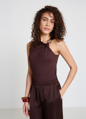 Donna con capelli ricci che indossa un top marrone scuro senza maniche con una fibbia decorativa e pantaloni abbinati a vita alta, con la mano in tasca.