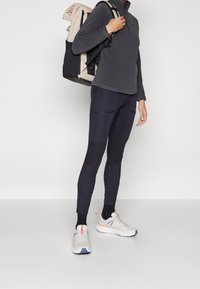 Persona vestida con camiseta gris oscuro, leggings negros con bolsillos con cremallera, zapatillas deportivas blancas y llevando una mochila beige y negra sobre el hombro.