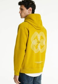 Ung mand i en sennepsgul hoodie med hvidt abstrakt design og tekst på ryggen, stående på siden mod en ensfarvet baggrund.