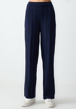 Marineblauwe wijde broek met verticale ribbeltextuur, elastische tailleband en ontspannen pasvorm, gecombineerd met witte casual sneakers.