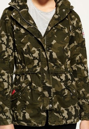 Osoba ubrana w zieloną i brązową kamuflażową kurtkę na zamek z przednimi kieszeniami i kapturem, pod którą ma białą i szarą pasiastą koszulę.