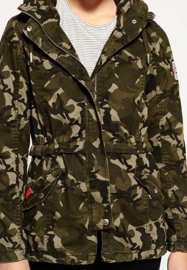 Personne portant une veste zippée à capuche avec un motif camouflage vert et marron, dotée de poches avant, sur une chemise rayée blanche et grise.