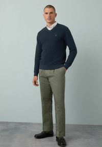 Marineblauwe geribbelde trui met V-hals, olijfgroene broek en zwarte schoenen. De outfit heeft een witte shirt met kraag eronder.