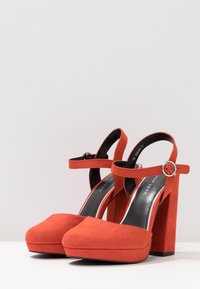 Pari di décolleté in suede rosso con plateau e cinturini alla caviglia, con fibbia argentata su superficie bianca contro uno sfondo grigio chiaro.