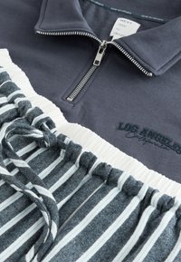 Szürke pulóver ezüst cipzárral, "LOS ANGELES" hímzéssel. Alatta csíkos farmer rövidnadrág, ráncolt derékrésszel és húzózsinórral.