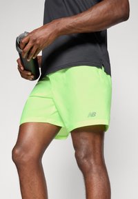 Athlet in neon grünen New Balance Shorts und einem schwarzen Shirt, hält eine schwarze Wasserflasche, Beine und Oberkörper sind sichtbar.