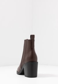 Brązowy skórzany botek sięgający kostki z teksturowanym wykończeniem po bokach, okrągłym noskiem i grubym obcasem. Posiada pętelkę z tyłu ułatwiającą zakładanie.