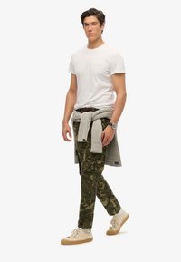 Witte t-shirt met korte mouwen, groene camouflage cargobroek, grijze trui om de taille geknoopt, beige sneakers en een polsbandhorloge.