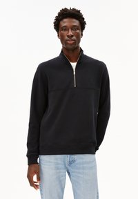 Schwarzer Reißverschluss-Sweatshirt mit hohem Kragen, mit einer horizontalen Naht über der Brust und gerippten Bündchen, getragen über einem weißen T-Shirt.