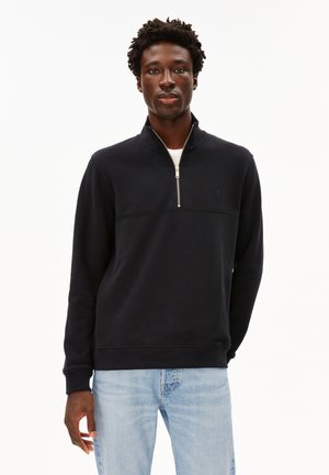 Schwarzer Reißverschluss-Sweatshirt mit hohem Kragen, mit einer horizontalen Naht über der Brust und gerippten Bündchen, getragen über einem weißen T-Shirt.