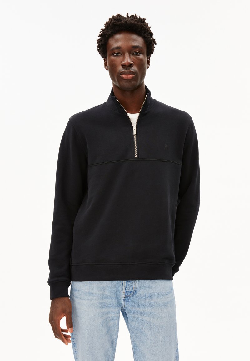 Schwarzer Reißverschluss-Sweatshirt mit hohem Kragen, mit einer horizontalen Naht über der Brust und gerippten Bündchen, getragen über einem weißen T-Shirt.