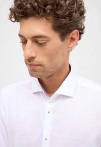 Witte shirt gemaakt van gestructureerde stof, met een kleurrijke, gemusterde kraagvoering en twee knopen aan de voorkant.