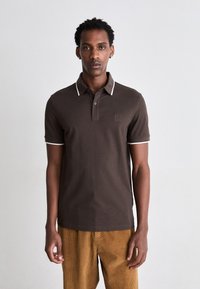 Braunes Poloshirt aus Baumwolle mit weißem Besatz am Kragen und an den Ärmeln. Verfügt über ein kleines quadratisches Logo auf der Brust. Die Cordhosen sind senfgelb.
