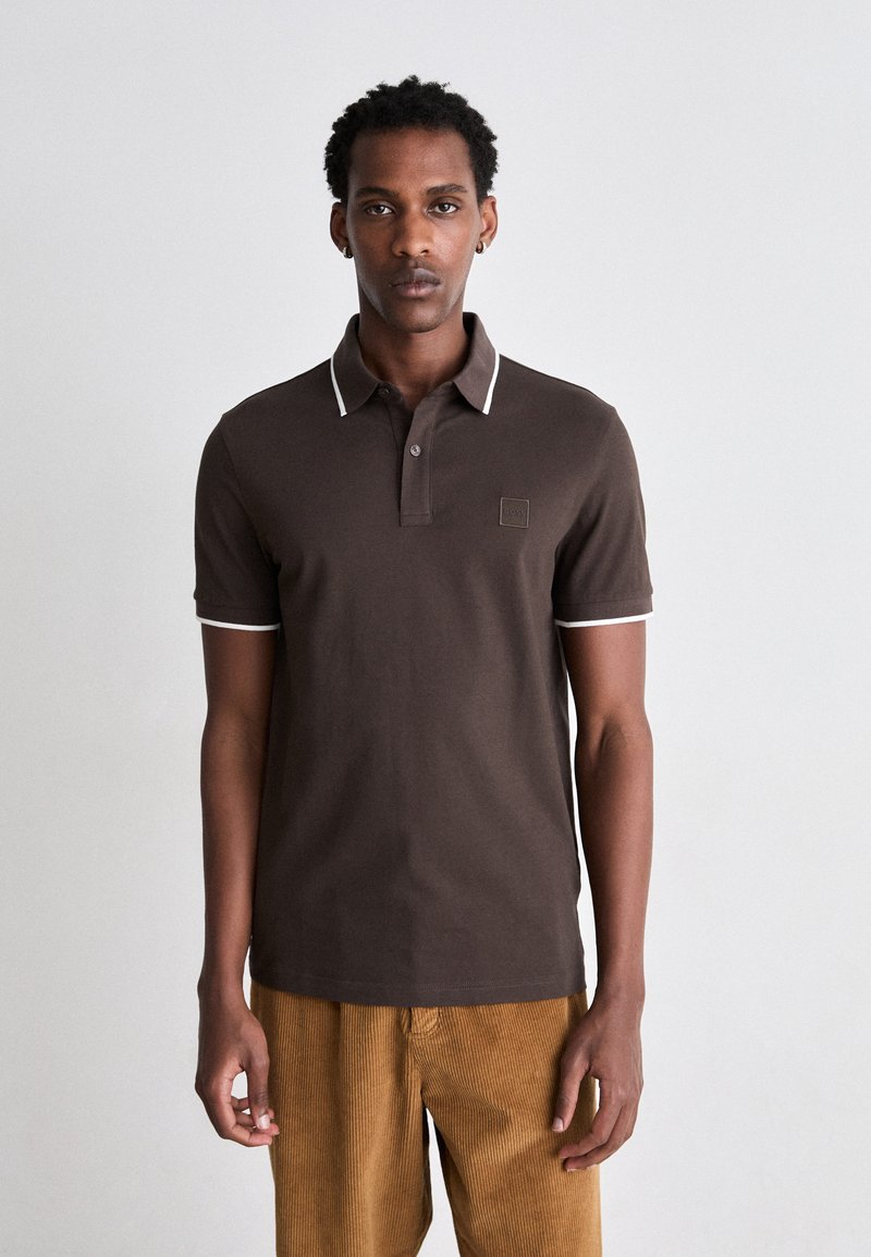 Braunes Poloshirt aus Baumwolle mit weißem Besatz am Kragen und an den Ärmeln. Verfügt über ein kleines quadratisches Logo auf der Brust. Die Cordhosen sind senfgelb.