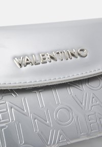 Pochette argentée avec un motif de texte en relief, une texture métallique lisse et une finition brillante. Logo en argent bien en vue.