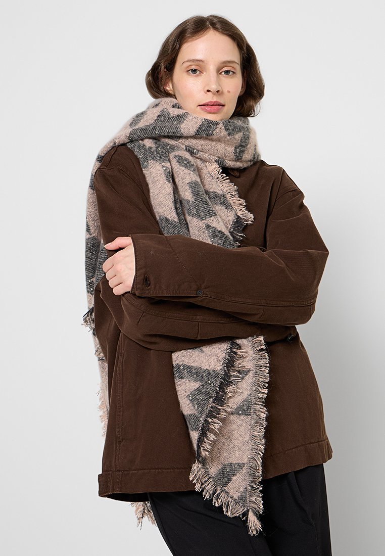 Mujer vestida con una chaqueta marrón y una gran bufanda de patrón beige y gris con flecos, de pie con los brazos cruzados frente a un fondo sencillo.