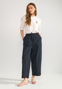 Femme aux longs cheveux portant une chemise blanche à manches courtes avec des motifs cœurs, un pantalon large noir et des chaussures roses à enfiler, debout les mains dans les poches.