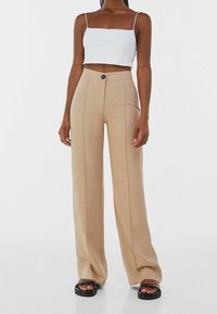 Helle beige, weit geschnittene Hose aus glattem Material, mit einem vorderen Knopfverschluss und vertikalen Nähten. Kombiniert mit einem weißen, cropped Oberteil.