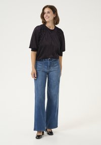 Top negro de manga corta con plisados y cuello redondo, combinado con jeans de mezclilla de pierna ancha de color azul claro y tacones negros, de pie contra un fondo blanco.