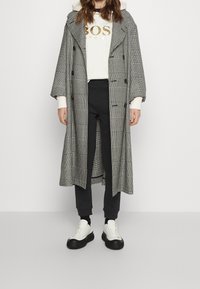 Kabát s černobílým houndstooth vzorem, dvouřadovým zapínáním, velkým límcem a oversized střihem, nošený přes bílou mikinu a černé kalhoty.