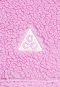 Rosa fleecetyg med en texturerad yta, som har en vit triangulär logotyp med "aCG" sydd i mitten. Mjuk och varm framtoning.