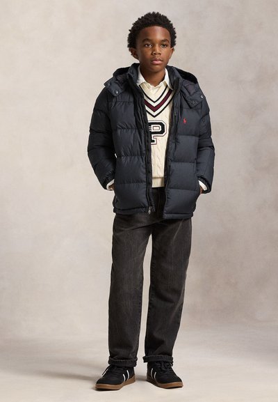 Chlapec stojí, nosí černou prošívanou bundu, krémový svetr s emblémem "P", bílou košili, tmavé džíny a černé tenisky s gumovou podrážkou.