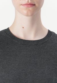 Gros plan sur le bas du visage et le cou d'une personne portant un pull en tricot gris foncé, avec un grain de beauté brun unique sur le côté gauche du cou.
