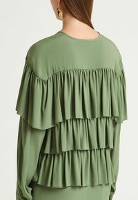 Grüne, geschichtete Bluse mit drei Rüschenlagen, weichem Stoff, Rundhalsausschnitt und glatter Textur. Schlankes Design mit fließender Silhouette.