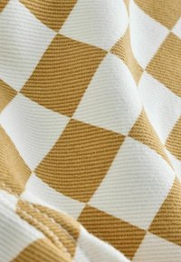 Texto textil con un patrón de cuadros que alterna entre cuadrados dorados y blancos. La tela tiene una apariencia texturizada, con costuras visibles a lo largo del borde.