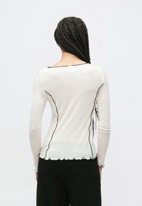 Bluza albă cu mâneci lungi, cu finisaj texturat și margini negre ondulate contrastante de-a lungul decolteului și tivului. Vedere din spate.