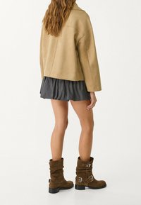 Femme aux cheveux longs et ondulés portant une veste marron clair, une mini-jupe plissée noire et des bottes en suède marron montant jusqu'au milieu des mollets avec des boucles, vue de dos.