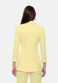 Blazer giallo su misura con silhouette aderente, maniche lunghe e polsini arrotolati, realizzato in tessuto liscio e caratterizzato da un design classico con revers.