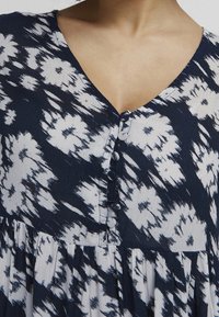 Námořnická a bílá vzorovaná blůza s výstřihem do V, s knoflíkovým zapínáním a detailním skladovým sukní, vyrobená z lehkého materiálu.