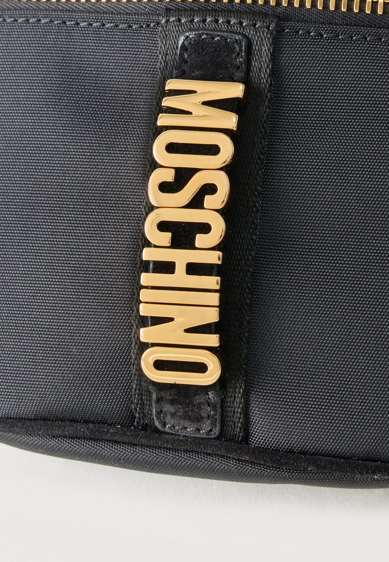 Black textured fabric with a gold vertical MOSCHINO logo attached near a visible gold zipper on a black accessory.