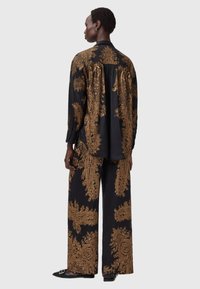 Inimene seisab seljaga, kandes musta ja kuldset paisley-mustriga pikkade varrukatega särki ja laiad püksid, jalas mustad terava ninaga kingad.