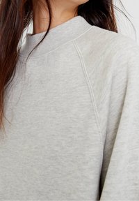 Sweat-shirt gris clair avec un col rond côtelé et des manches raglan. Tissu doux et texturé, présentant des nuances subtiles pour plus de détails.