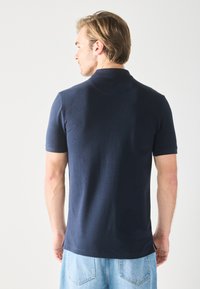Polo shirt marine en tissu lisse, avec un col et des manches courtes. Porté avec un short en denim bleu clair. Vue de dos, design simple.