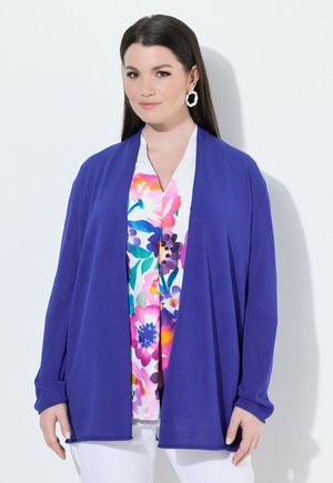 Kvinde med langt brunt hår bærer hvide øreringe, en lilla åben cardigan, en farverig blomsterbluse og hvide bukser, stående mod en hvid baggrund.