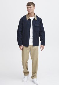 Marinblå jacka med en ljusbrun krage, framdragkedja och fyra fickor; ihop med beige byxor och vita sneakers.