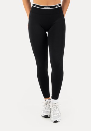 Ben iført sorte tætsiddende atletikleggings med et mærket taljebånd og hvide sneakers med hvide ankelstrømper på en lys baggrund.