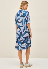 Hellblaues Kleid mit abstrakten Blättern in Dunkelblau und Rot, kurzen Ärmeln, schichted Rock und einem Kragen. Getragen mit beigen Turnschuhen.