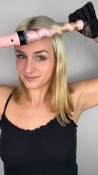 Fer à boucler avec un barrel rose ondulé et une base noire tenu près de cheveux blonds ; l'utilisateur porte un débardeur noir et un gant noir à la main.
