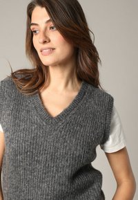Gilet rayé en tricot gris avec un décolleté en V, porté par-dessus une chemise blanche. Texture douce, design sans manches et détails subtils sur le tissu.