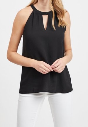 Kvinde iført en sort ærmeløs halterneck med en frontnøgleudskæring og hvide bukser, hænderne løst foldet foran.