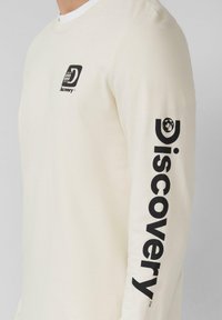 Langsleeve shirt van crème stof met een zwart Discovery-logo op de borst en grote zwarte tekst langs de linkermouw.