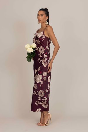 Roșie închisă, rochie din satin cu flori, cu bretele subțiri, design lung până la podea, cu modele mari de flori crem și roz, purtată cu sandale cu curele.