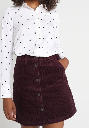 Woman wearing a white long-sleeve shirt with black stars and a buttoned dark purple corduroy skirt with pockets.