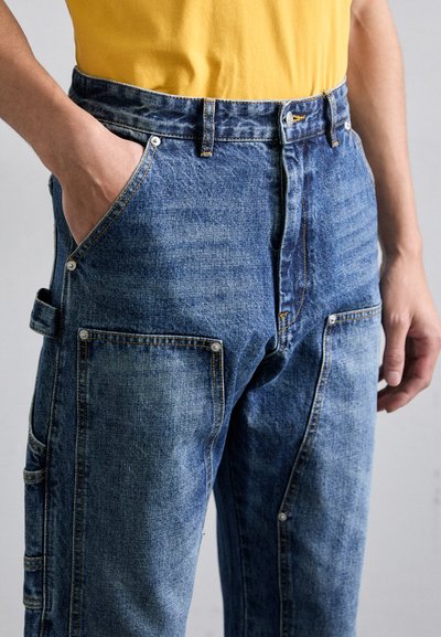 Un pantalon cargo en denim de bleu moyen avec une coupe décontractée, cinq poches et des détails à boutons-pression à l'avant et sur le côté.