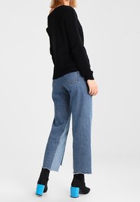 Pull noir côtelé, jean bleu taille haute avec un ourlet brut, et bottines noires à talons carrés turquoise, vus de dos.