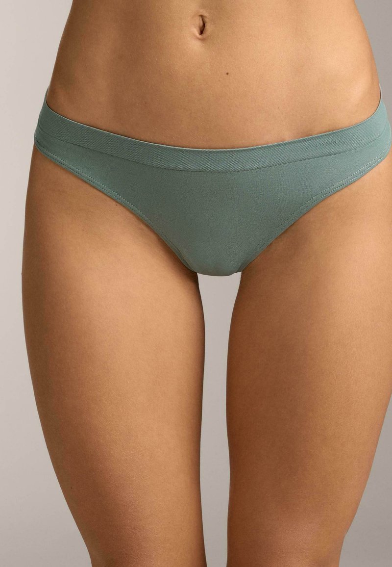 Femme portant un sous-vêtement bikini sans coutures vert clair, montrant la taille, les hanches et le haut des cuisses sur un fond neutre.